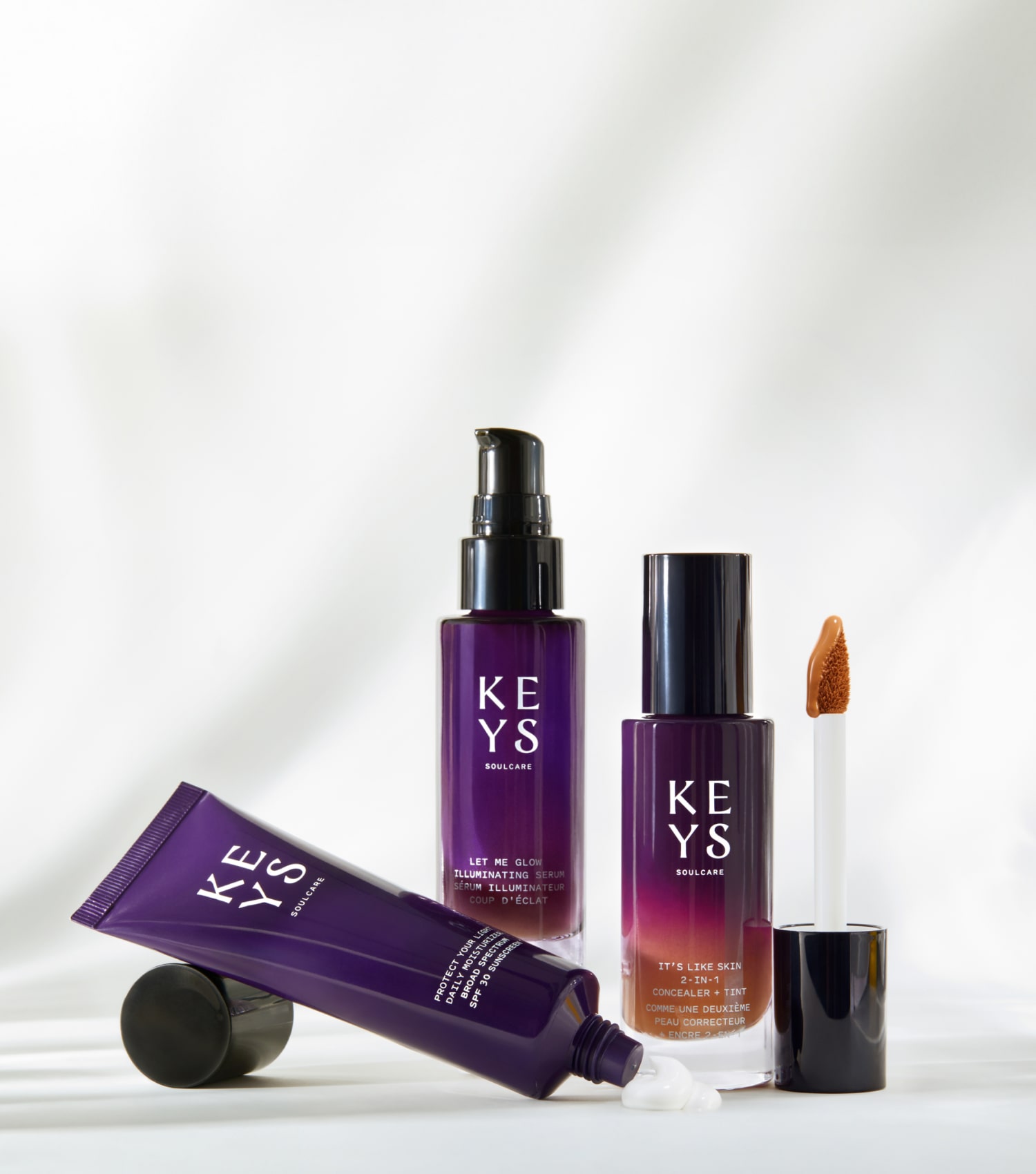 Promotional image featuring a collection of 'KEYS Soulcare' beauty products elegantly arranged against a white backdrop with flowing lines suggesting draped fabric. The assortment includes a purple squeeze tube, a pump bottle, and a concealer with its applicator wand visible, all with the brand's logo in white against the purple packaging. The text 'SHOP ALL' is prominent on the right side, in large uppercase letters, followed by the invitation 'Explore skincare, makeup, and body care rituals, created by Alicia Keys.
