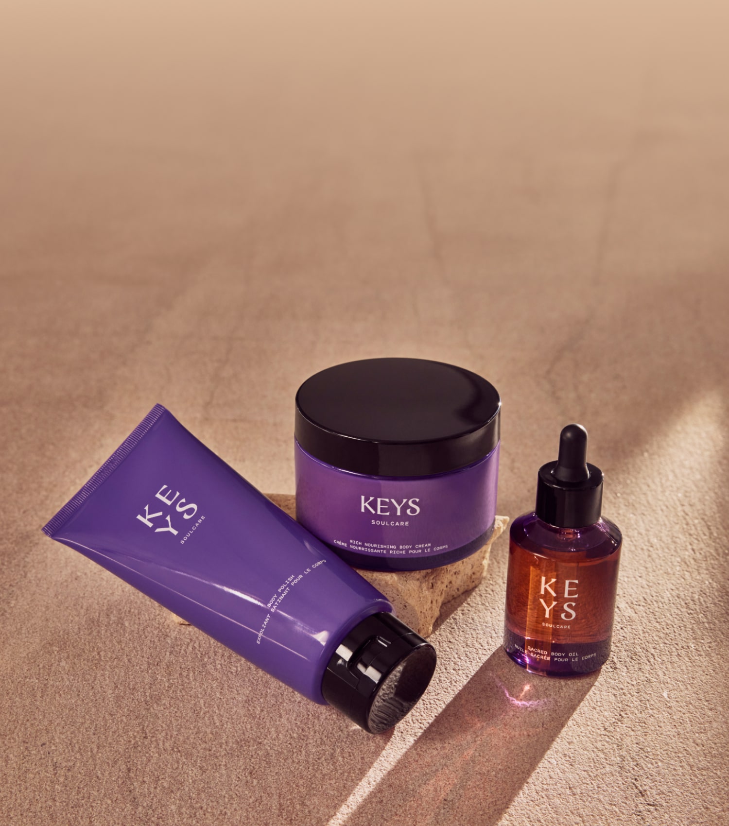 Promotional banner displaying a collection of body care products from the 'KEYS Soulcare' line. The products are arranged on a textured beige surface that resembles soft fabric, bathed in warm light that creates a relaxed ambiance. From left to right, there is a purple squeeze tube, possibly for body wash, a large black-lidded jar labeled 'Nourishing Body Cream', and a smaller purple glass dropper bottle likely containing body oil. The text 'BODY CARE' is in large, uppercase black letters, with a tagline underneath that reads 'Care for your whole self with body wash, body oil, tools, and more.