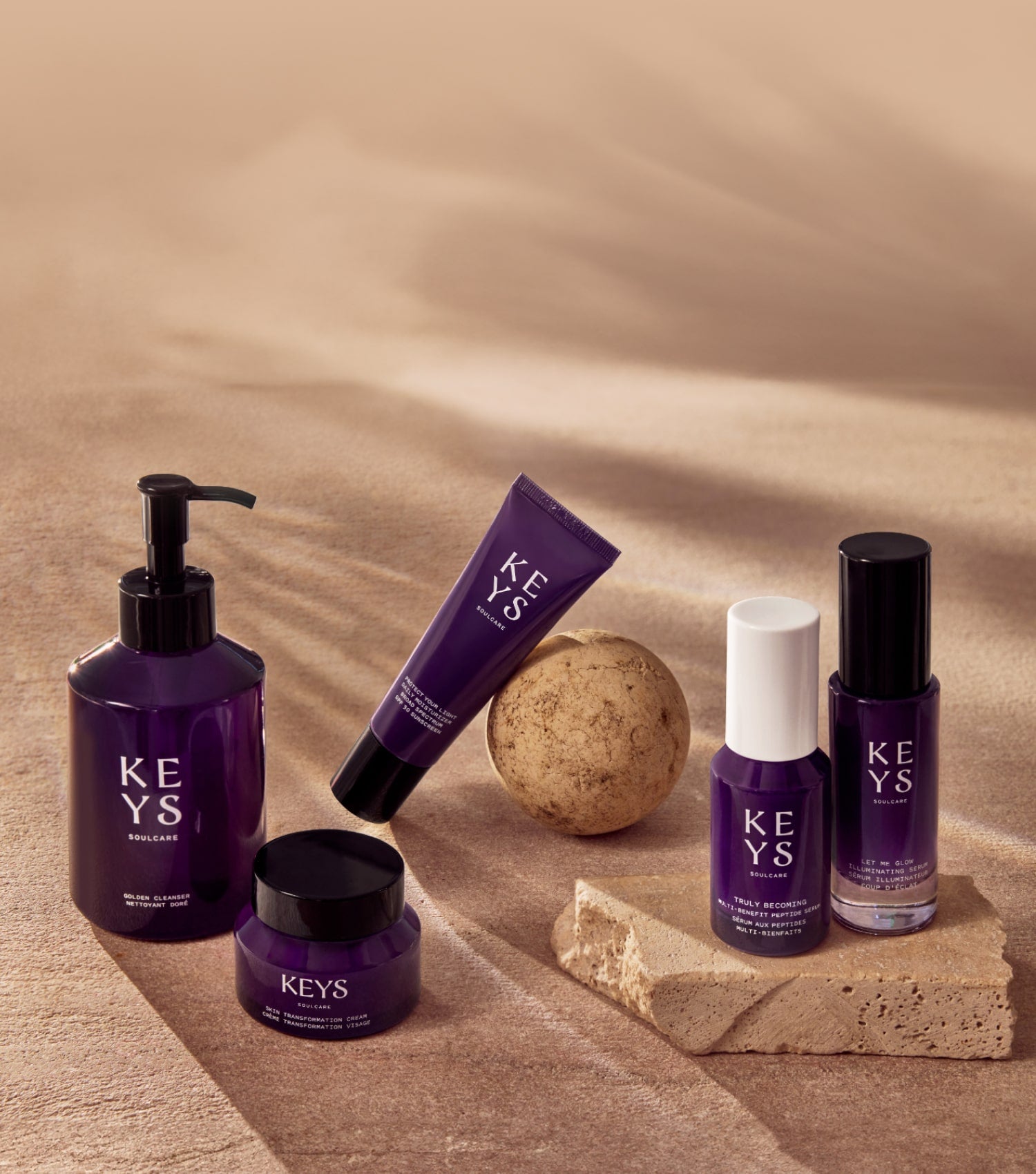 Elegant promotional image for a 'KEYS Soulcare' best sellers collection. The products are displayed on a beige, sandy background with a natural texture. Included in the arrangement is a lit candle in a black container, a small jar of cream, a glass dropper bottle, a pump dispenser bottle, and a small spray bottle, all in a cohesive dark purple color scheme. The large text 'BEST SELLERS' is seen to the right, in an uppercase font, complemented by a message in smaller letters that reads 'Turn your makeup and skincare routines into rituals with these most-loved essentials.