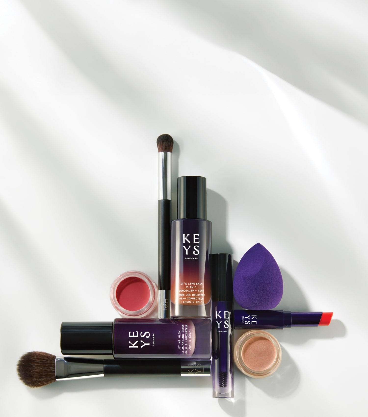Promotional image featuring an array of makeup products arranged on a white surface with shadows suggesting soft lighting. The products include a makeup brush, a bottle of foundation, a mascara wand, a red lipstick, a purple makeup sponge, and a small open jar of cosmetic cream. The text 'MAKEUP' is prominently displayed in large black letters, followed by a smaller caption 'Discover skincare-makeup hybrids for a natural-looking glow.' The products are marked with the brand 'KEYS' in capitalized white text on purple packaging.