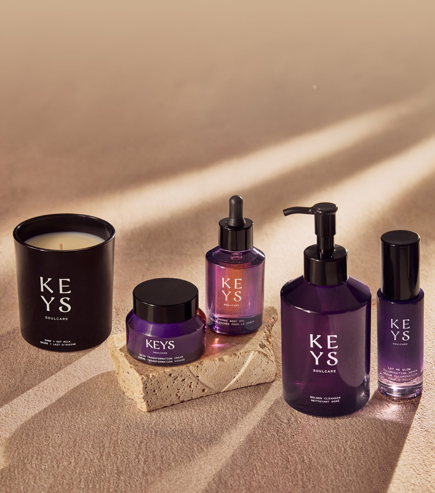 Elegant promotional image for a 'KEYS Soulcare' best sellers collection. The products are displayed on a beige, sandy background with a natural texture. Included in the arrangement is a lit candle in a black container, a small jar of cream, a glass dropper bottle, a pump dispenser bottle, and a small spray bottle, all in a cohesive dark purple color scheme. The large text 'BEST SELLERS' is seen to the right, in an uppercase font, complemented by a message in smaller letters that reads 'Turn your makeup and skincare routines into rituals with these most-loved essentials.